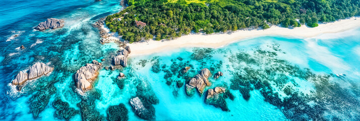 Aerial view of tropical island coastline with turquoise ocean water, white sandy beach, granite rock formations, and dense green forest vegetation in Indian Ocean