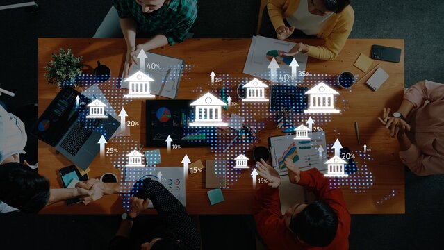 A top-down view of a diverse team engaged in a business meeting, analyzing financial trends and data with charts, laptops, and digital representations on a wooden table. Raster