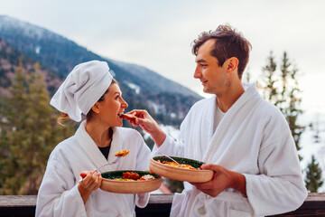 Couple ordered breakfast in hotel room eating food on balcony wearing robes enjoying winter mountains landscape.