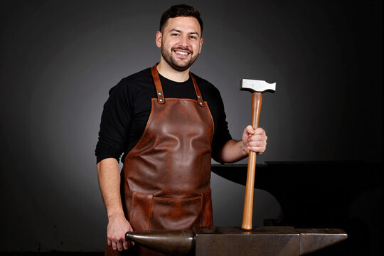 Happy modern blacksmith or craftsman in a leather apron, smiling at the camera while holding a hammer over an anvil in his workshop, representing traditional craft and skilled manual labor