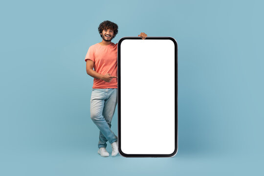 A stylish indian man with curly hair and a beard is posing next to a large blank smartphone screen. He is pointing at the empty display and smiling.