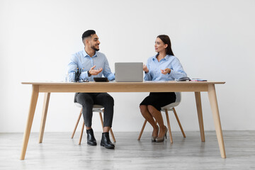 Obraz premium Two colleagues, a smiling woman and a middle eastern man, are seated at a desk with a laptop. They are engaged in a friendly discussion about their business project in a contemporary office.