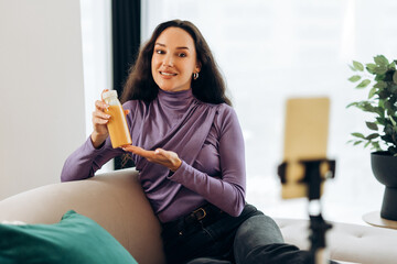 Happy woman showing bottle with smoothie recording video, sitting on comfortable sofa in living room
