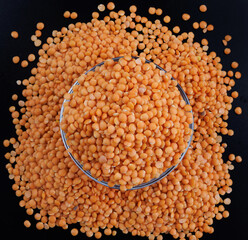 Red lentils in a transparent glass cup on a black background.. A pile of lentils around the cup.