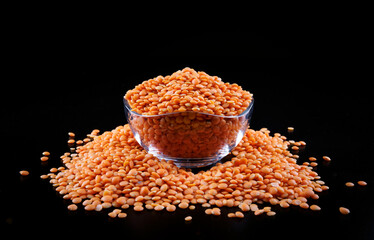Red lentils in a transparent glass cup on a black background. Side view. A pile of lentils around the cup.