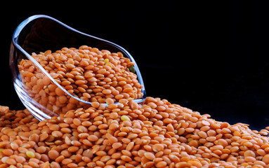 Red lentils in a transparent glass cup on a black background. Side view. Lots of lentils around the cup.