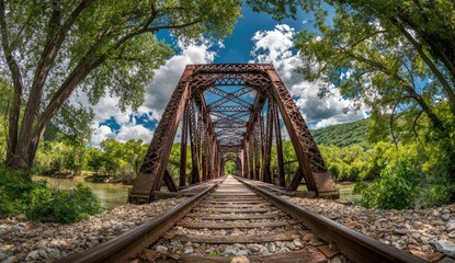 Old railway bridge spans a river through lush green trees under a bright sky.