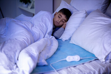 Little boy sleeping under blanket on electric heating pad in bedroom at night