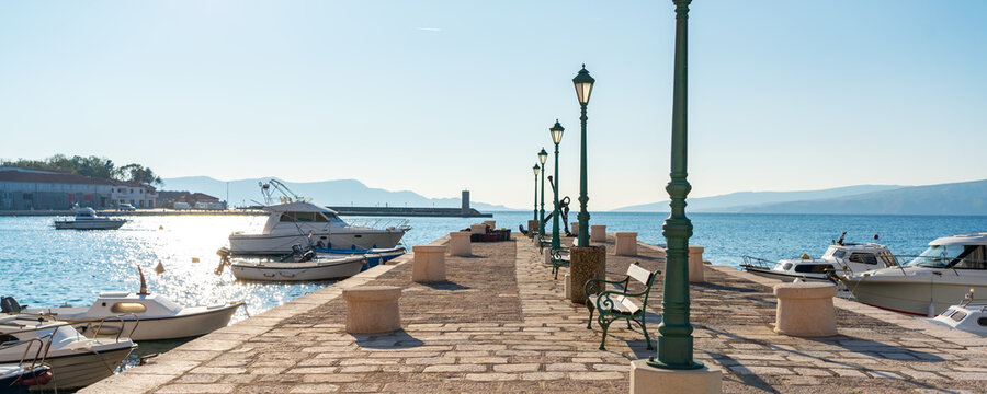 Fototapeta Scenic stone pier with moored boats and street lamps under clear blue sky in Senj, Croatia