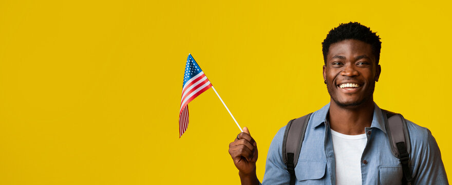 A man is proudly holding an American flag while smiling. He wears a light blue shirt and has a backpack on. The bright yellow background adds to the cheerful atmosphere of the scene.