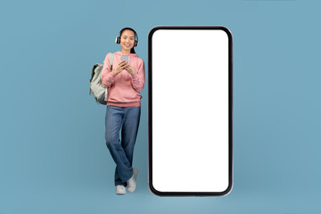 A cheerful young asian woman with a headset and smartphone stands next to a huge mobile phone with an empty screen. She is using an entertaining mobile app, showcasing a relaxed and happy moment.