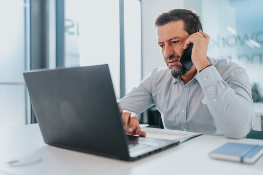 A focused professional multitasking with laptop and phone in a bright, contemporary office setting.