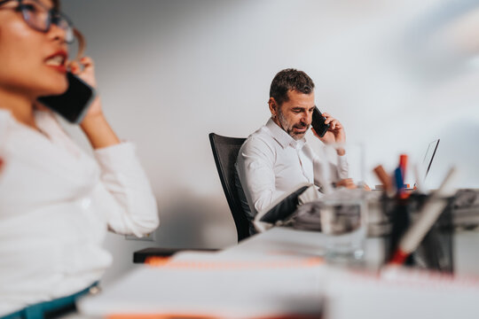 Two colleagues in a bright office use smart phones and laptops, conveying busy, connected work life.