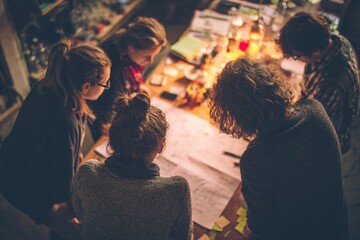Fototapeta premium NGO volunteers around a table planning a project, photographed from behind, natural warm tones. Teamwork in non profit organization. Volunteer work. Feel good business activity.