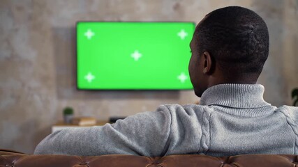 Rear view of an African American man sitting comfortably on a sofa intently watching a green screen television with tracking markers in a dimly lit living room perfect for content replacement.