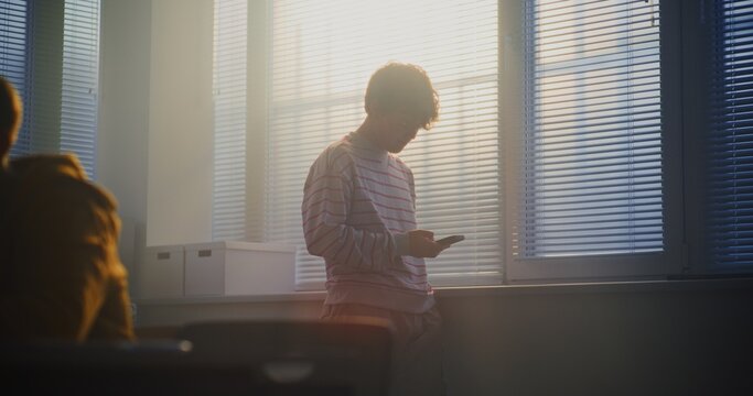 Male Student in Striped Sweatshirt Stands Near Bright Classroom Window, Checking Messages on Smartphone During Break. Concept Highlights Digital Habits, Youth Communication, Modern Student Lifestyle.