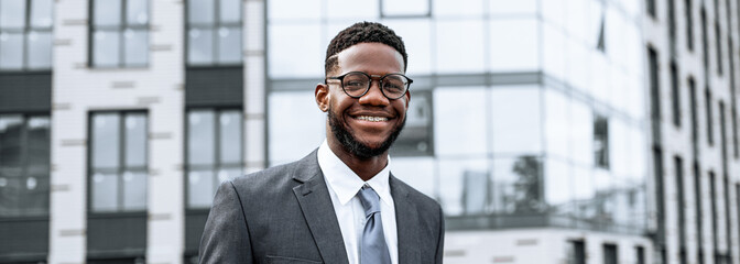 A young man dressed in a formal suit stands outside a contemporary building with glass windows. He...