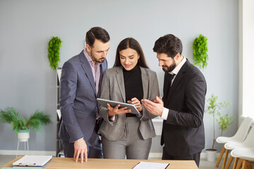 Group of smiling business people standing near table and discussing project on digital tablet. Two men and one woman in formal suits analyzing company strategy and financial data in office together.