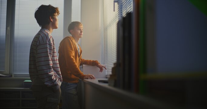 Two Teenage Boys Stand Together Near Sunlit Classroom Window, Talking Softly While Taking Short Break From Studies. Concept Casual Friendship, Relaxed Communication, Everyday School Life. Slow motion.