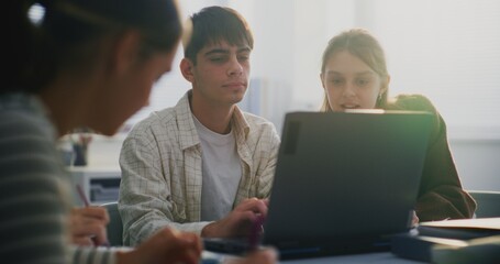 Young Male Student in Plaid Shirt Centered, Actively Talking and Pointing Laptop Screen While Collaborating With Two Female Classmates. Concept Active Peer Learning, Academic Discussion. Static Shot.