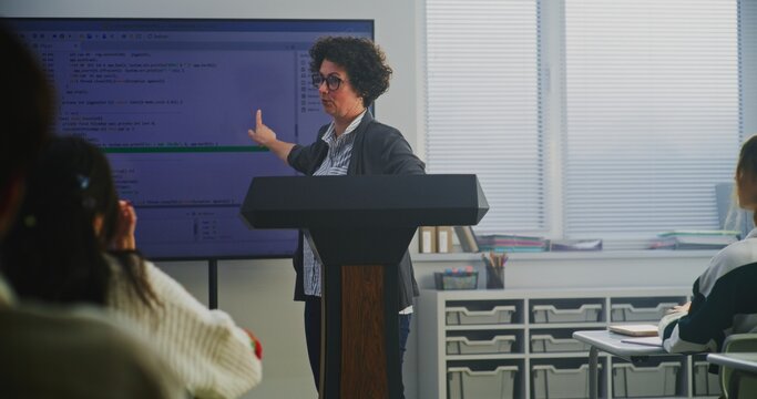 Female Coding Teacher Stands at Podium, Explaining Programming Syntax on Large Digital Screen. Concept Modern Computer Science Education, STEM Learning, Technology Literacy, and Teaching Innovation.