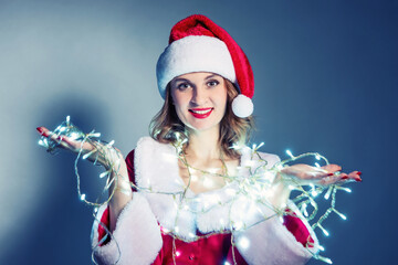 Smiling woman in Santa costume holding glowing LED Christmas lights, festive holiday portrait on blue background, cheerful winter celebration concept
