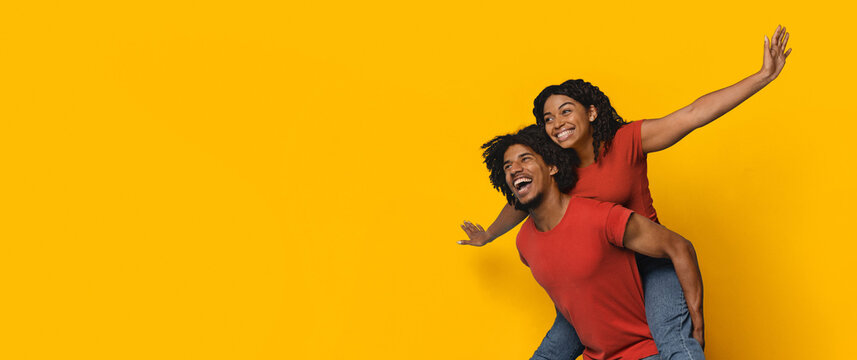 A happy couple is having fun, with one person giving a piggyback ride while smiling brightly. The lively yellow backdrop adds to the cheerful atmosphere of the scene.