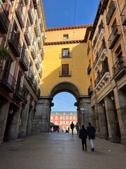 Historic stone archway framing Plaza Mayor with red buildings and spire under blue sky in Madrid, Spain