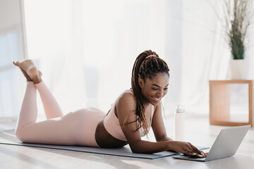 Fit young black woman in sportswear is lying on a mat, using her laptop to check fitness videos and relax after an intense home workout session. She enjoys a moment of rest and focus.