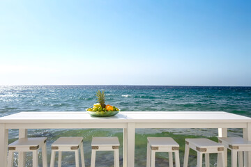 white wooden table with empty chairs on seascape backdrop under clear blue sky on sunny summer day. plate of fruit