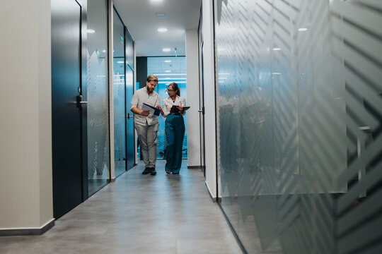 Two coworkers walk through a glass-walled corridor, discussing reports and planning next steps.