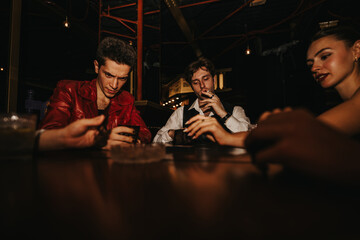 Three friends unwind at a dimly lit bar, sharing drinks, hands of cards, and conversation. A...