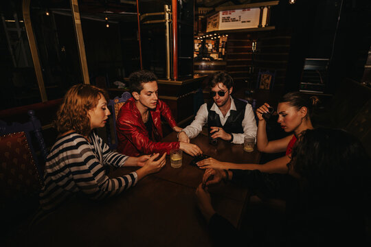 A group of five friends sits around a dark wooden table in a moody bar, enjoying drinks, conversation, and a card game, capturing a lively, social night out.