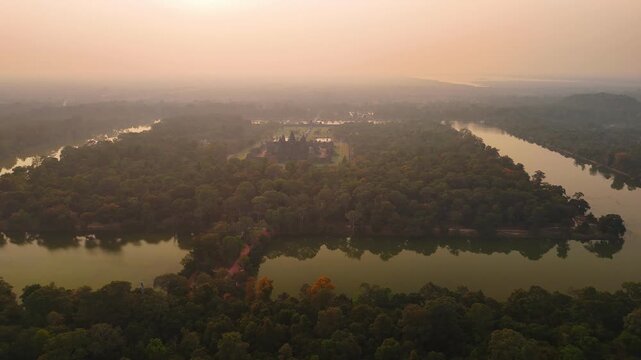 Aerial view of angkor wat temple complex surrounded by a moat and lush jungle during a warm, hazy sunrise in siem reap, Cambodia.
