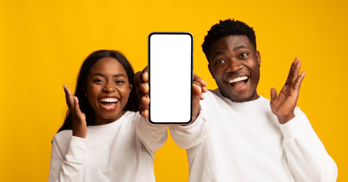 A joyful couple poses against a vibrant yellow backdrop, smiling widely while holding up a smartphone. They seem delighted as they share a moment of enthusiasm and connection.