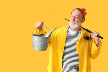 Senior man with fishing rod and caught fish in bucket on yellow background
