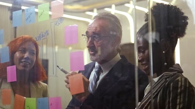 Diverse business team in modern office brainstorms as senior manager writes on glass with sticky notes. Corporate meeting showcases teamwork, collaboration, leadership, engaged colleagues. 4k video
