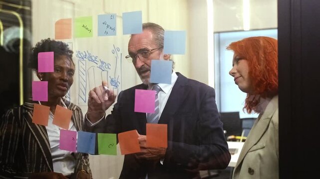 Diverse business team in modern office brainstorms as senior manager writes on glass with sticky notes. Corporate meeting showcases teamwork, collaboration, leadership, engaged colleagues. 4k video - Powered by Adobe