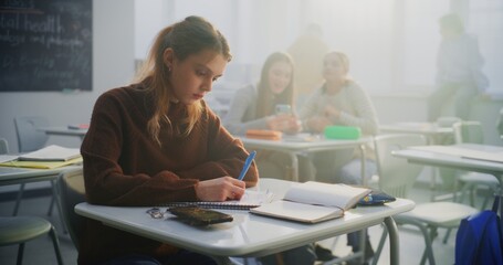 Sad Teenage Girl Sits Alone at Desk, Looking Down Notebook While Classmates Laugh and Chat in...