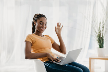 Lovely young black lady sits in a chair at home, wearing earphones and making a video call on her laptop. She waves at the webcam, smiling and communicating happily.