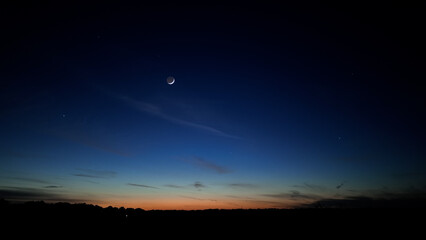 Blue hour time with crescent Moon, stars and planets above landscape silhouettes.