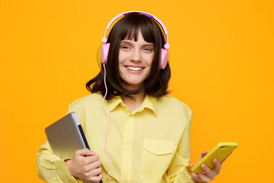 A cheerful young woman wearing pink headphones and a yellow shirt smiles at the camera, holding a tablet and smartphone against a bright orange backdrop for a vivid tech lifestyle portrait.