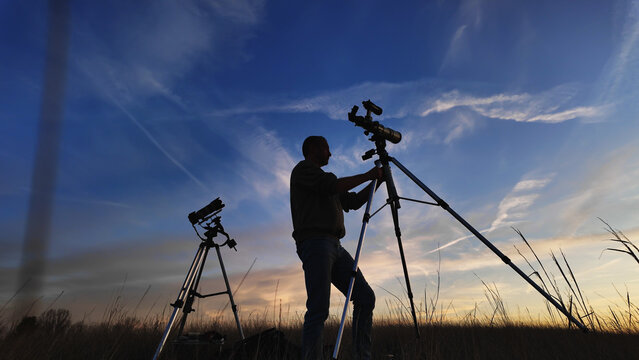 Amateur astro-photographer with a telescope and camera on a tripod photographing night sky with stars, planets, Moon and other celestial objects.