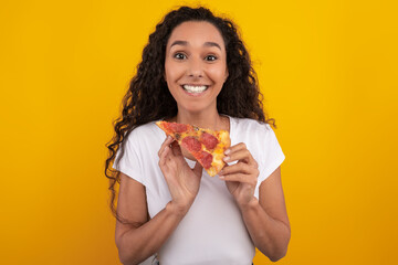 A happy millennial woman with curly hair stands in a studio against a yellow-orange background. She joyfully holds a pepperoni pizza slice and poses for the camera.