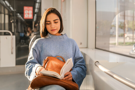 Young commuter woman sitting by window and reading book on public transportation - Powered by Adobe