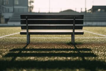 Empty wooden bench on a sunlit sports field at golden hour, long shadows across dewy turf with stadium seats in soft bokeh
