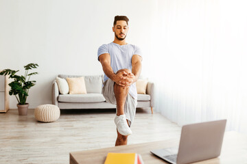 A young man is performing a yoga pose at home, balancing on one leg. He follows a video on his laptop in a bright, airy living room with plants and comfortable seating.