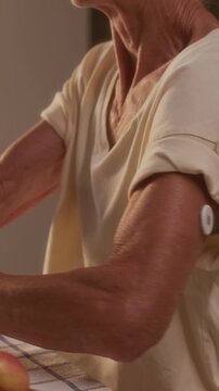 Medium vertical of elderly woman with continuous glucose monitoring patch sitting in kitchen after having lunch, testing blood sugar with smartphone app and getting red low reading