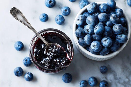 Overhead close-up of ripe blueberries in a white bowl and glossy homemade blueberry jam in a jar with a vintage spoon on marble surface