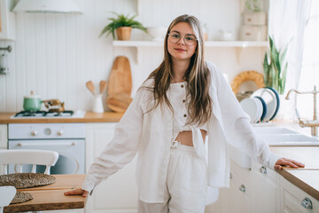 A woman in white loungewear stands confidently in a brightly lit, stylish kitchen, blending casual elegance with domestic charm.